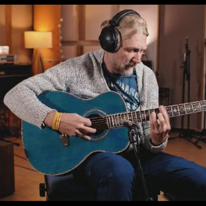 A man wearing over-ear headphones sits and plays a Byron OM Body Full-Size Left-Handed Guitar Ocean Blue in a warmly lit music studio or room with instruments and equipment visible in the background.