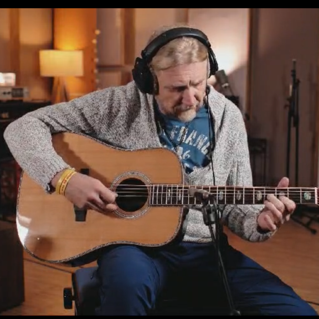 A man wearing headphones sits in a music studio, playing an acoustic guitar. The studio has warm lighting, wooden floors, and musical instruments in the background.